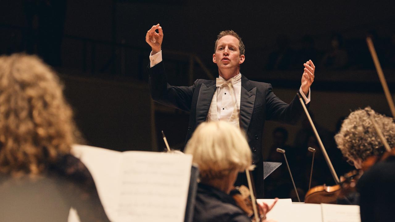 Justin Doyle dirigert das Neujahrskonzert vom RIAS Kammerchor und hebt dabei beide für die Musiker von der Akademie für Alte Musik Berlin die Arme.