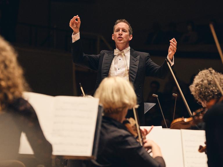 Justin Doyle dirigert das Neujahrskonzert vom RIAS Kammerchor und hebt dabei beide für die Musiker von der Akademie für Alte Musik Berlin die Arme.