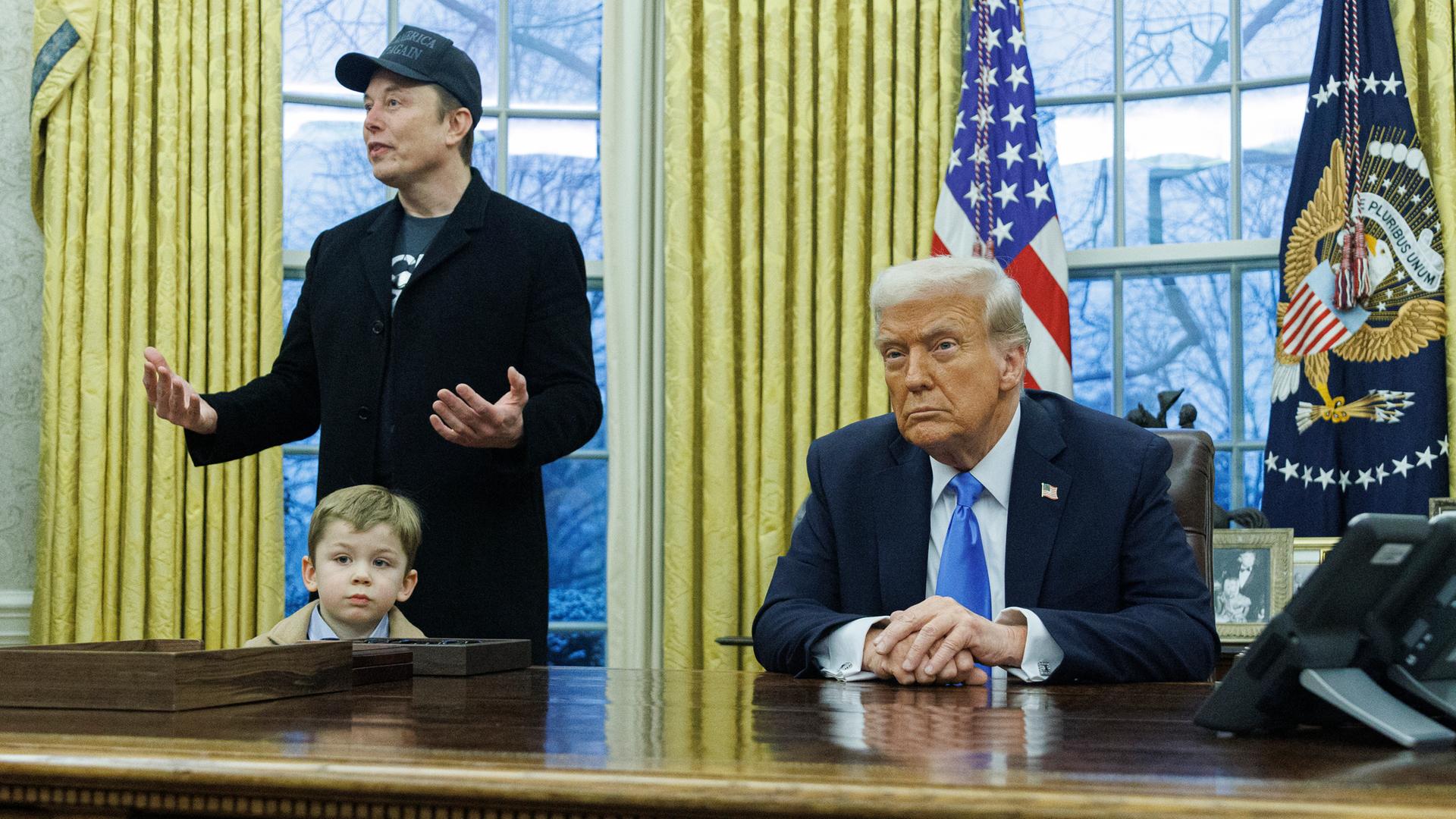 Elon Musk erklärt etwas bei einer Pressekonferenz im Oval Office, während sein vierjähriger Sohn X Æ A-Xii vor seinen Beinen steht. Am Schreibtisch sitzt Donald Trump und schaut konzentriert in die Kamera.