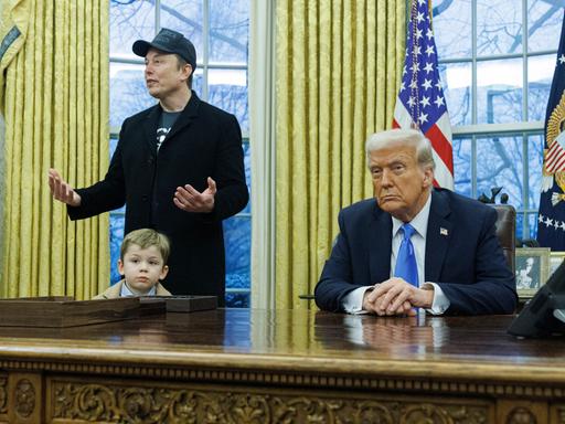 Elon Musk erklärt etwas bei einer Pressekonferenz im Oval Office, während sein vierjähriger Sohn X Æ A-Xii vor seinen Beinen steht. Am Schreibtisch sitzt Donald Trump und schaut konzentriert in die Kamera.