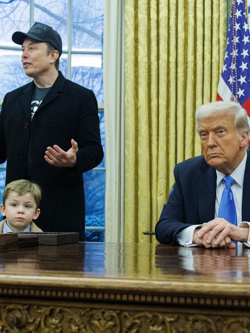 Elon Musk erklärt etwas bei einer Pressekonferenz im Oval Office, während sein vierjähriger Sohn X Æ A-Xii vor seinen Beinen steht. Am Schreibtisch sitzt Donald Trump und schaut konzentriert in die Kamera.