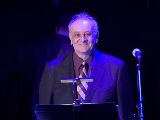 Der Komponist Angelo Badalamenti steht in Anzug und Krawatte an einem Notenpult.