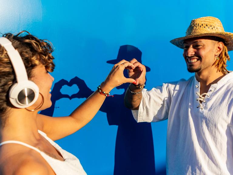 Ein Mann mit Dreadlocks und eine Frau mit Kopfhörer formen mit ihren Händen gemeinsam ein Herz vor einem blauen Hintergrund.