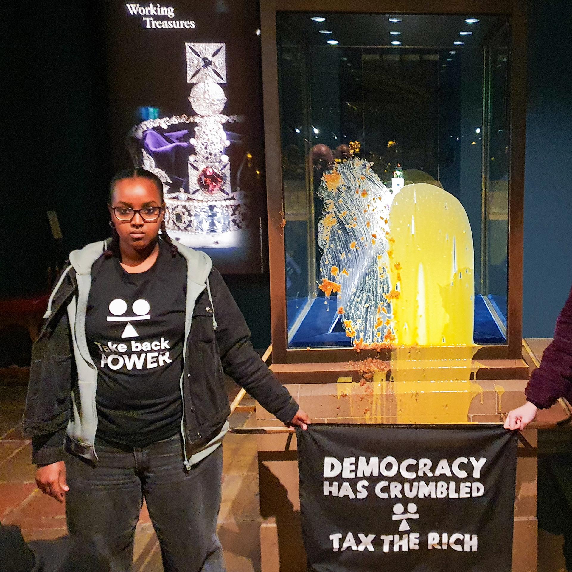 Aktivisten im Tower of London nach einer "Lebensmittelattacke" auf die Kronjuwelen - sie haben Shirts mit der Aufschrift "Take back Power" an und halten ein Transparent in den Händen auf dem steht "Tax the rich" - also "Besteuer die Reichen". 