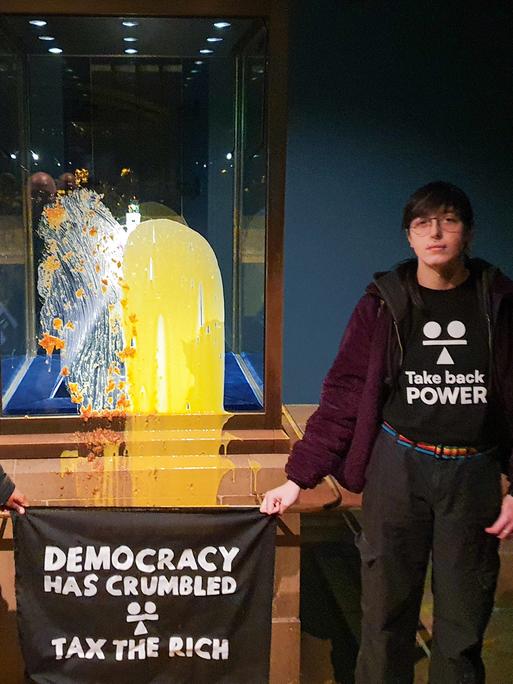 Aktivisten im Tower of London nach einer "Lebensmittelattacke" auf die Kronjuwelen - sie haben Shirts mit der Aufschrift "Take back Power" an und halten ein Transparent in den Händen auf dem steht "Tax the rich" - also "Besteuer die Reichen". 