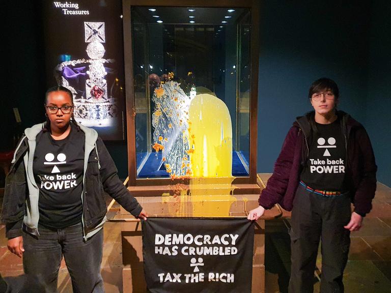 Aktivisten im Tower of London nach einer "Lebensmittelattacke" auf die Kronjuwelen - sie haben Shirts mit der Aufschrift "Take back Power" an und halten ein Transparent in den Händen auf dem steht "Tax the rich" - also "Besteuer die Reichen". 