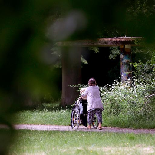 Eine ältere Dame schiebt eine Person in einem Rollstuhl durch einen Park. 