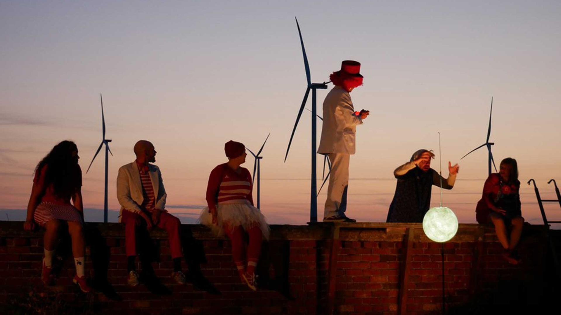 Schauspieler agieren auf und hinter einer Mauer mit einer weißen Mondlaterne, am Abendhimmel sind Windräder zu sehen.