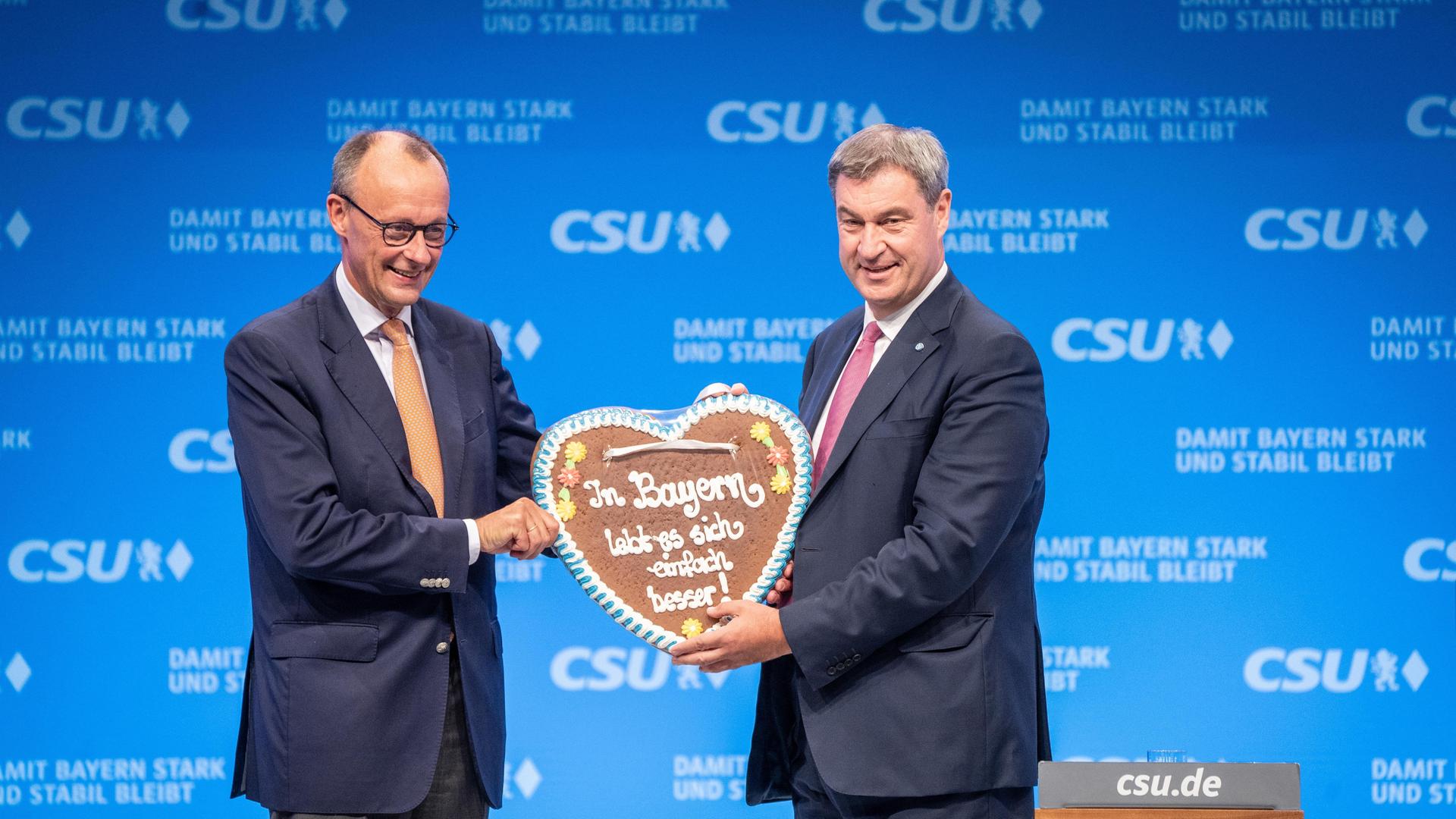 Auf dem CSU-Parteitag 2023 in München übergibt Markus Söder Friedrich Merz ein Lebkuchenherz mit der Aufschrift "In Bayern lebt es sich einfach besser!".