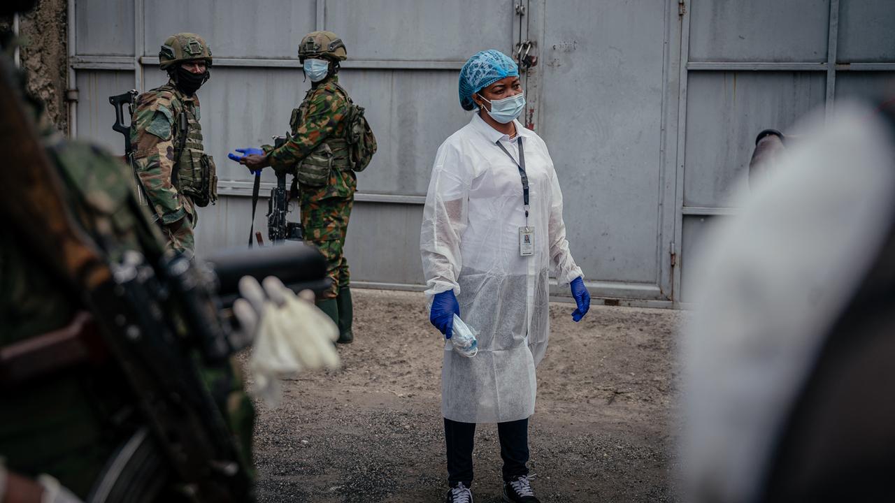 Eine Person in medizinischer Kleidung und Mund- und Nasenschutz steht zwischen Soldaten, die ebenfalls einen Mund- und Nasenschutz tragen. Eine Person in medizinischer Kleidung und Mund- und Nasenschutz steht zwischen Soldaten, die ebenfalls einen Mund- und Nasenschutz tragen.