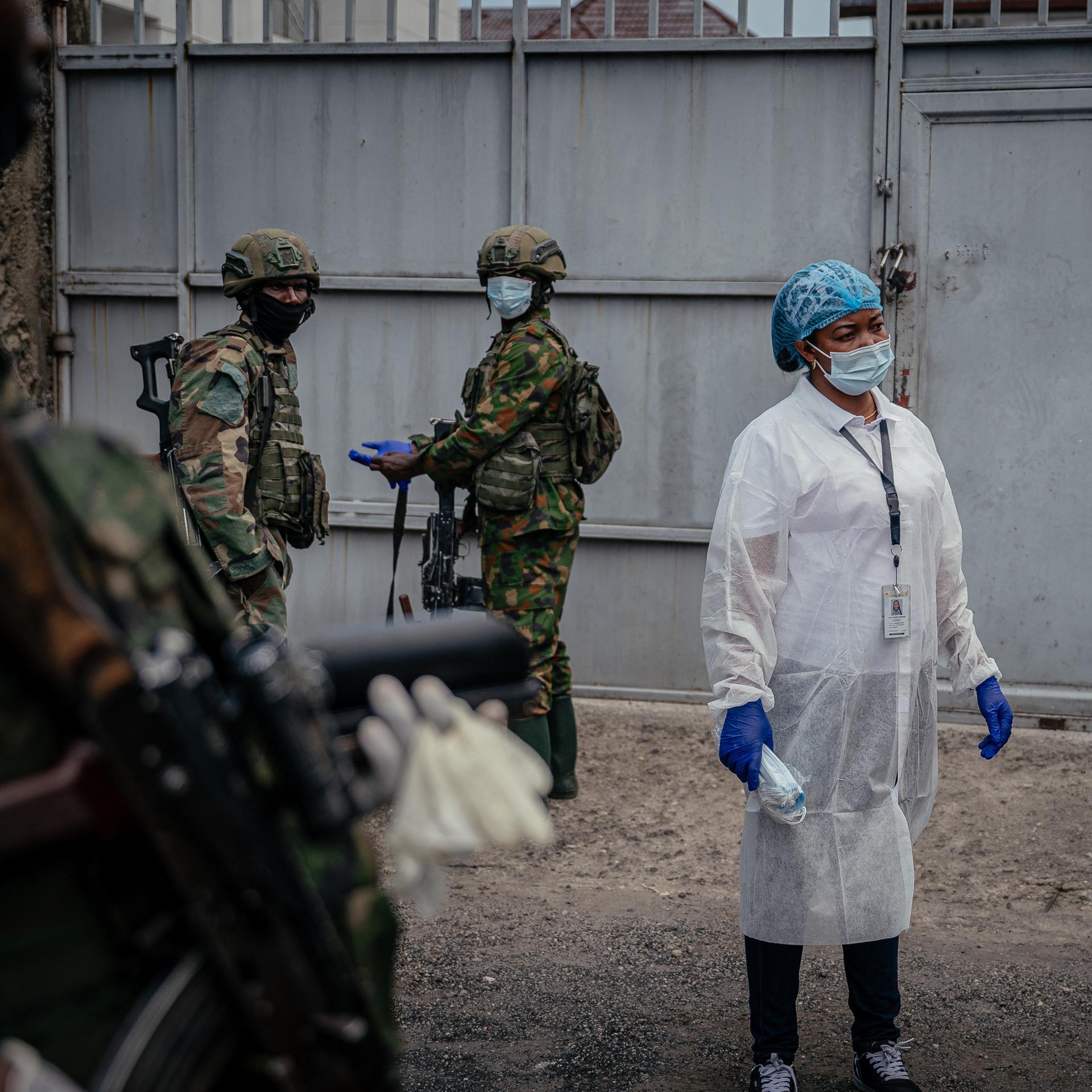 Eine Person in medizinischer Kleidung und Mund- und Nasenschutz steht zwischen Soldaten, die ebenfalls einen Mund- und Nasenschutz tragen.