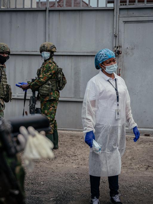 Eine Person in medizinischer Kleidung und Mund- und Nasenschutz steht zwischen Soldaten, die ebenfalls einen Mund- und Nasenschutz tragen. Eine Person in medizinischer Kleidung und Mund- und Nasenschutz steht zwischen Soldaten, die ebenfalls einen Mund- und Nasenschutz tragen.