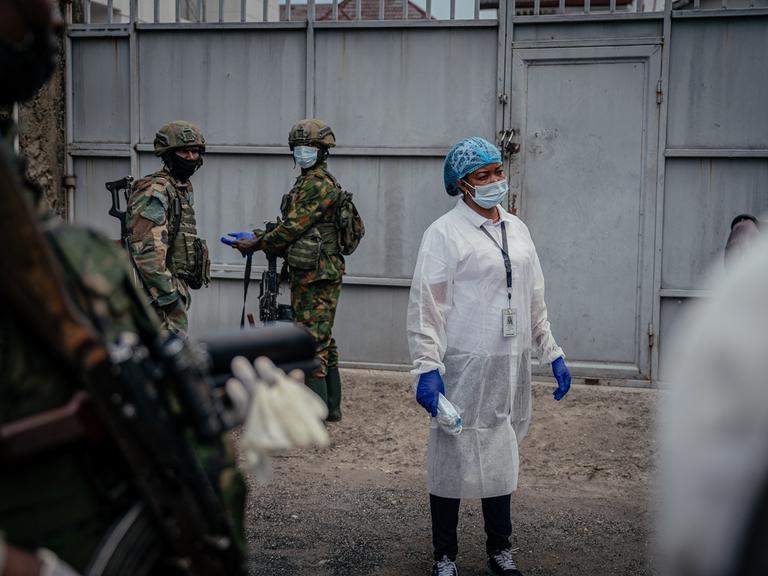 Eine Person in medizinischer Kleidung und Mund- und Nasenschutz steht zwischen Soldaten, die ebenfalls einen Mund- und Nasenschutz tragen. Eine Person in medizinischer Kleidung und Mund- und Nasenschutz steht zwischen Soldaten, die ebenfalls einen Mund- und Nasenschutz tragen.