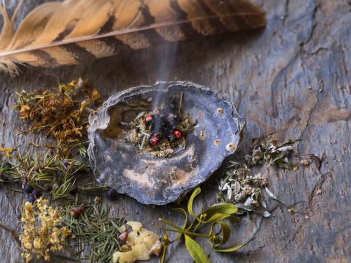 Auf einer Steinplatte liegen getrocknete und zerkleinerte Kräuter für die Rauhnachts-Räucherung. Aus einer Muschelschale steigt leichter Rauch auf, oberhalb ist eine Feder. Auf einer Steinplatte liegen getrocknete und zerkleinerte Kräuter für die Rauhnachts-Räucherung. Aus einer Muschelschale steigt leichter Rauch auf, oberhalb ist eine Feder.