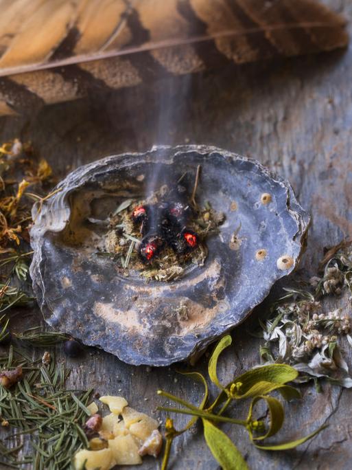 Auf einer Steinplatte liegen getrocknete und zerkleinerte Kräuter für die Rauhnachts-Räucherung. Aus einer Muschelschale steigt leichter Rauch auf, oberhalb ist eine Feder.