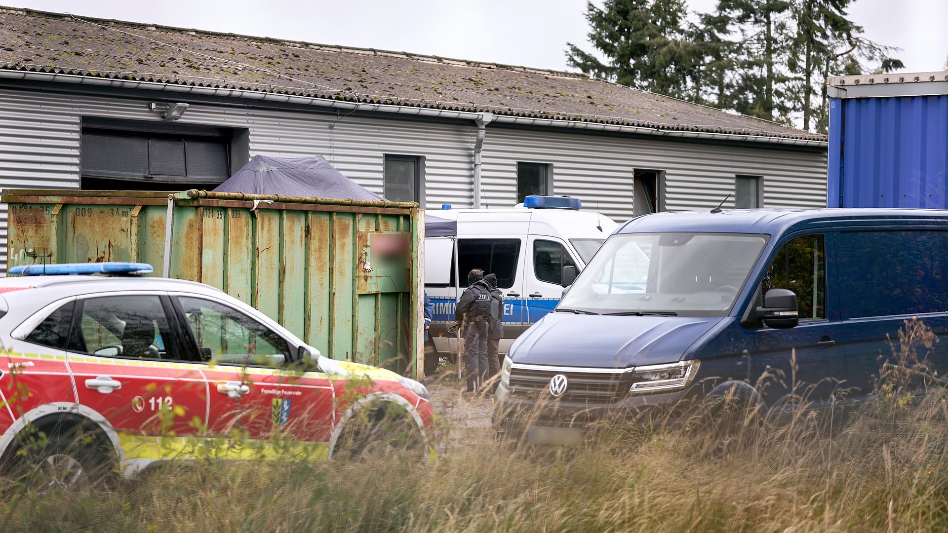 Vor einer Halle und einem Container stehen Fahrzeuge der Feierwehr, der Polizei und des Technischen Hilfswerks. Im Vordergrund hochgewachsenes Gras. 