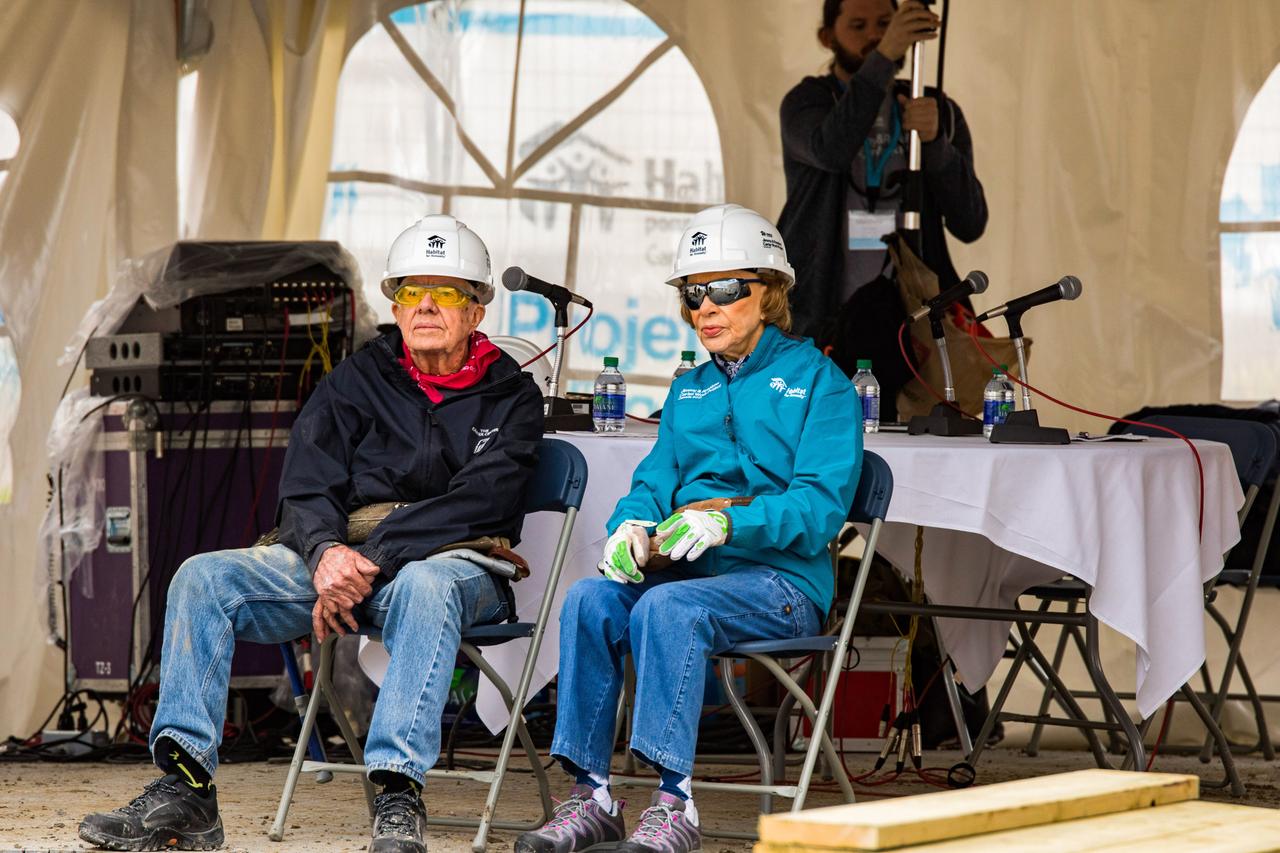 Jimmy und Rosalynn Carter sitzen in Arbeitskleidung auf Klappstühlen. Sie tragen Bauhelme und Schutzbrillen.