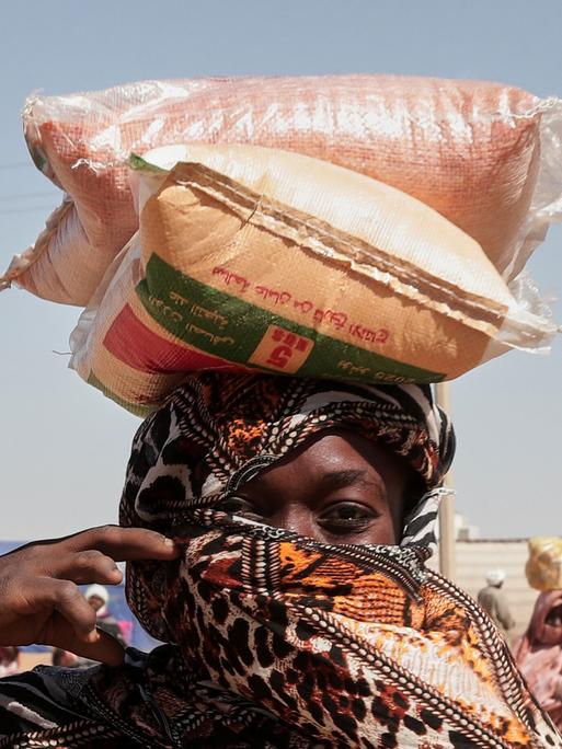 Eine sudanesische Geflüchtete mit Kopftuch trägt Säcke mit Nahrungsmitteln auf dem Kopf Eine sudanesische Geflüchtete mit Kopftuch trägt Säcke mit Nahrungsmitteln auf dem Kopf