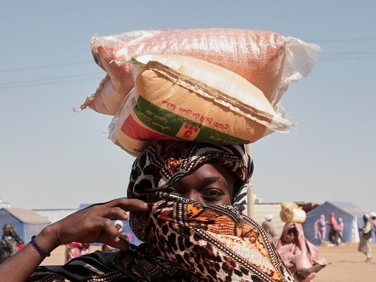 Eine sudanesische Geflüchtete mit Kopftuch trägt Säcke mit Nahrungsmitteln auf dem Kopf Eine sudanesische Geflüchtete mit Kopftuch trägt Säcke mit Nahrungsmitteln auf dem Kopf