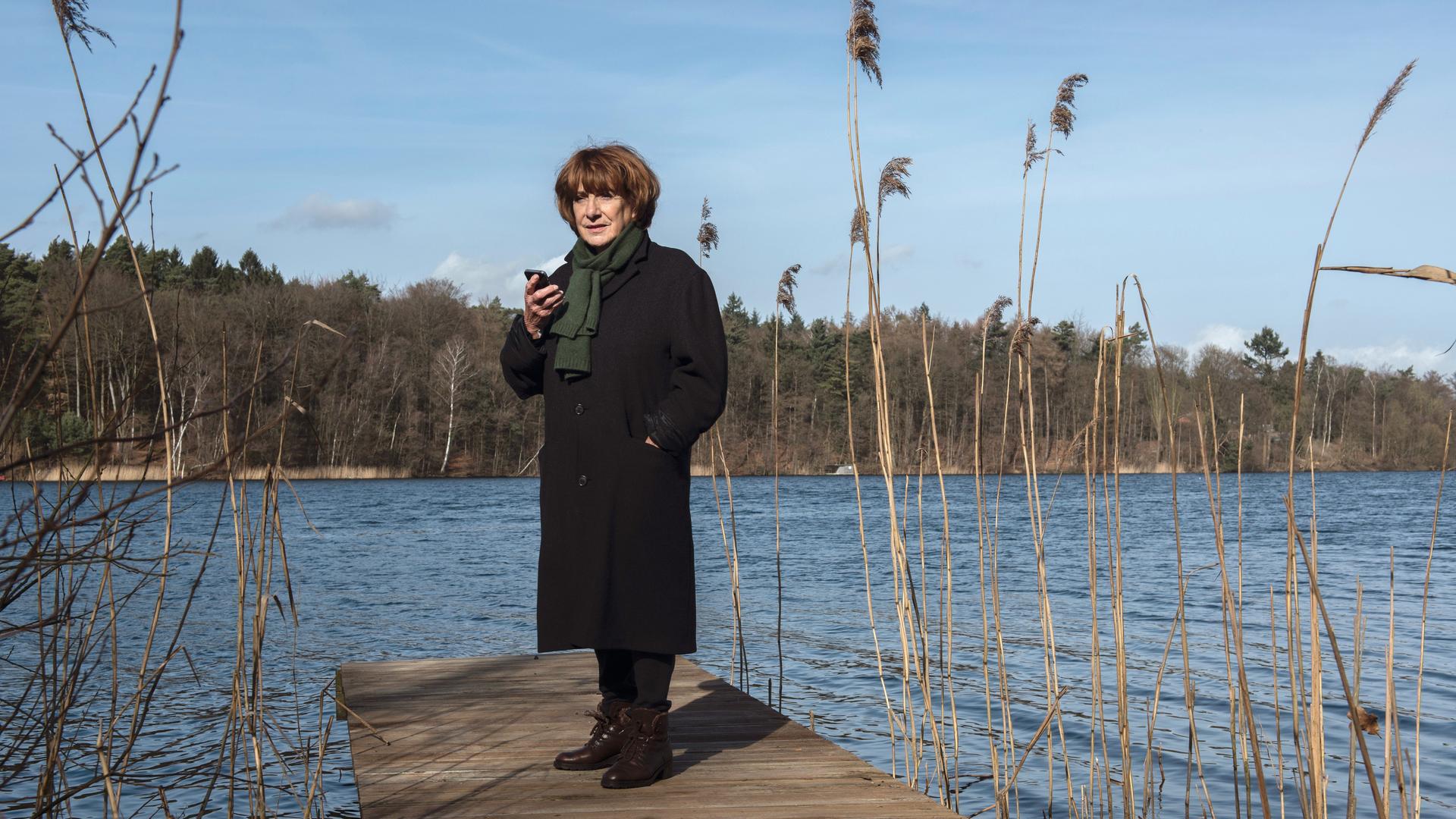 Im Still aus "Bella Block" steht Hannelore Hoger in einem schwarzen Mantel auf einem Steg, hinter ihr ist ein See zu sehen.