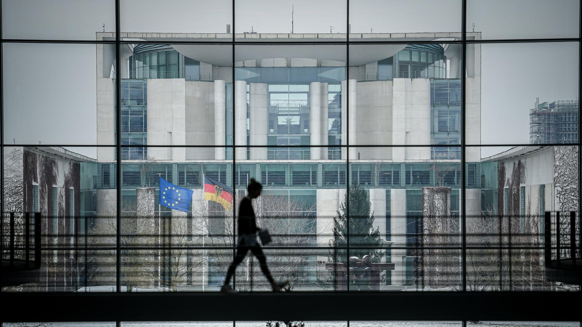 Blick vom Innern des Kanzleramtes nach außen. Im Vordergrund geht vor einer großen Glasfront eine Person über einen Flur. Durch die Glasfront sieht man Außenbereiche des Gebäudes.