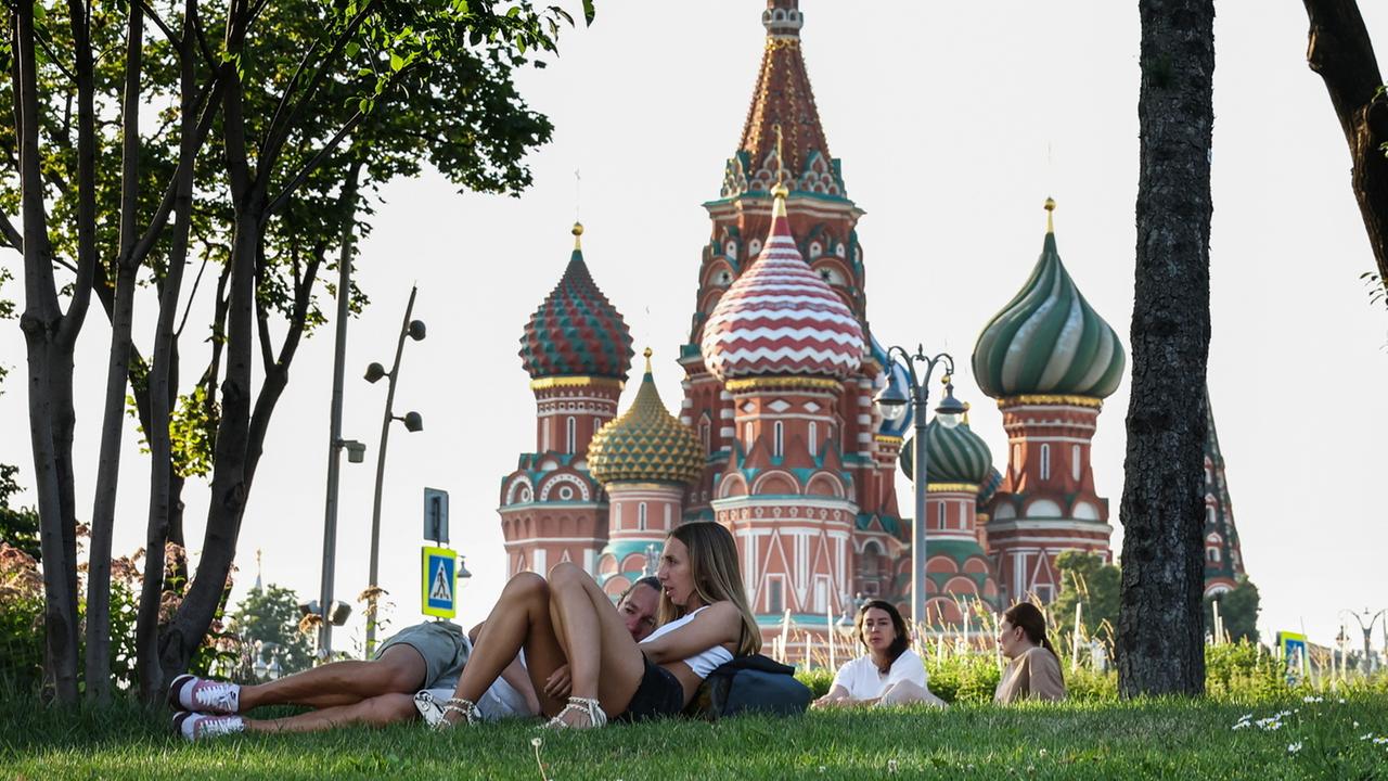 Junge Menschen liegen auf einer Wiese in einem Park, im Hintergrund ist eine russische Kirche mit Zwiebeltürmen zu sehen.