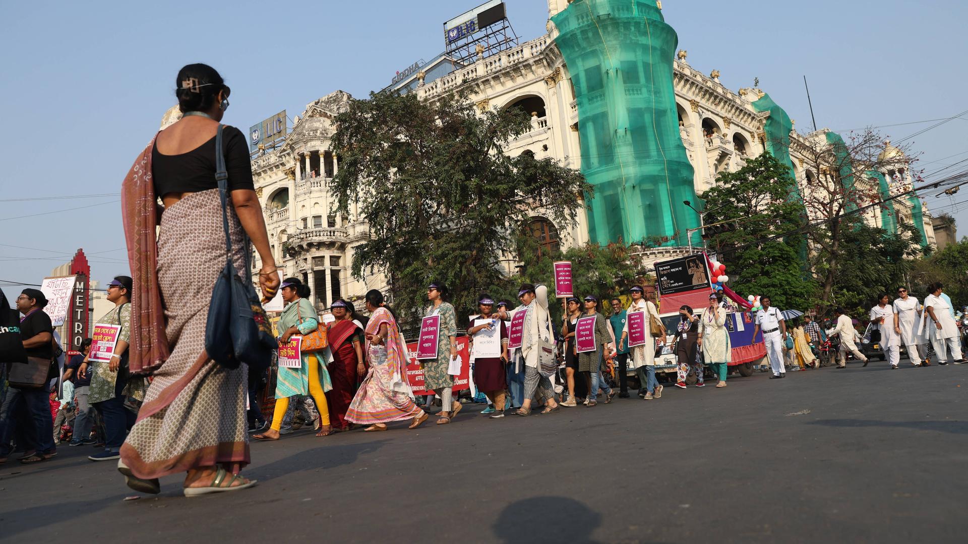 Auf einer Straße in Kolkata marschieren Personen mit Transparenten. Im Vordergrund geht eine Frau in die entgegengesetzte Richtung.