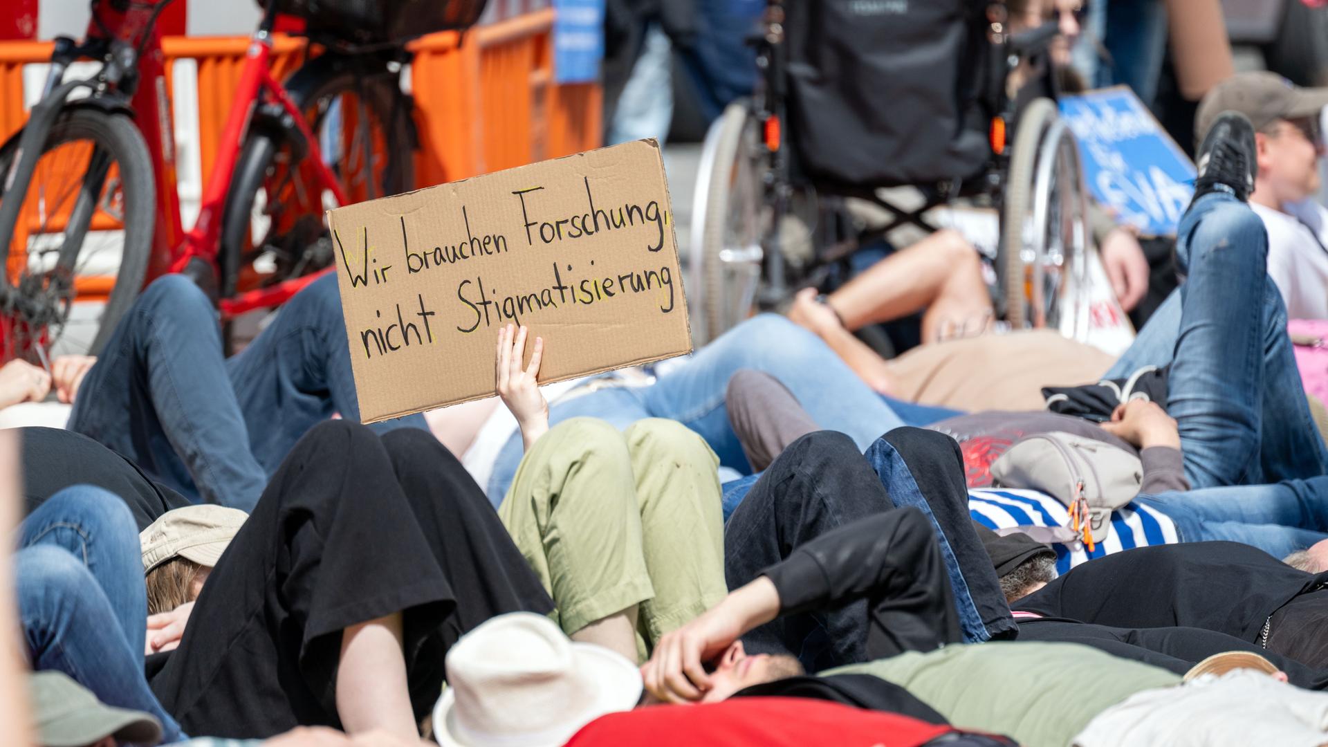 Zahlreiche Menschen liegen nebeneinander auf der Straße, während eine Frau ein Schild mit der Aufschrift "Wir brauchen Forschung, nicht Stigmatisierung" hoch hält.