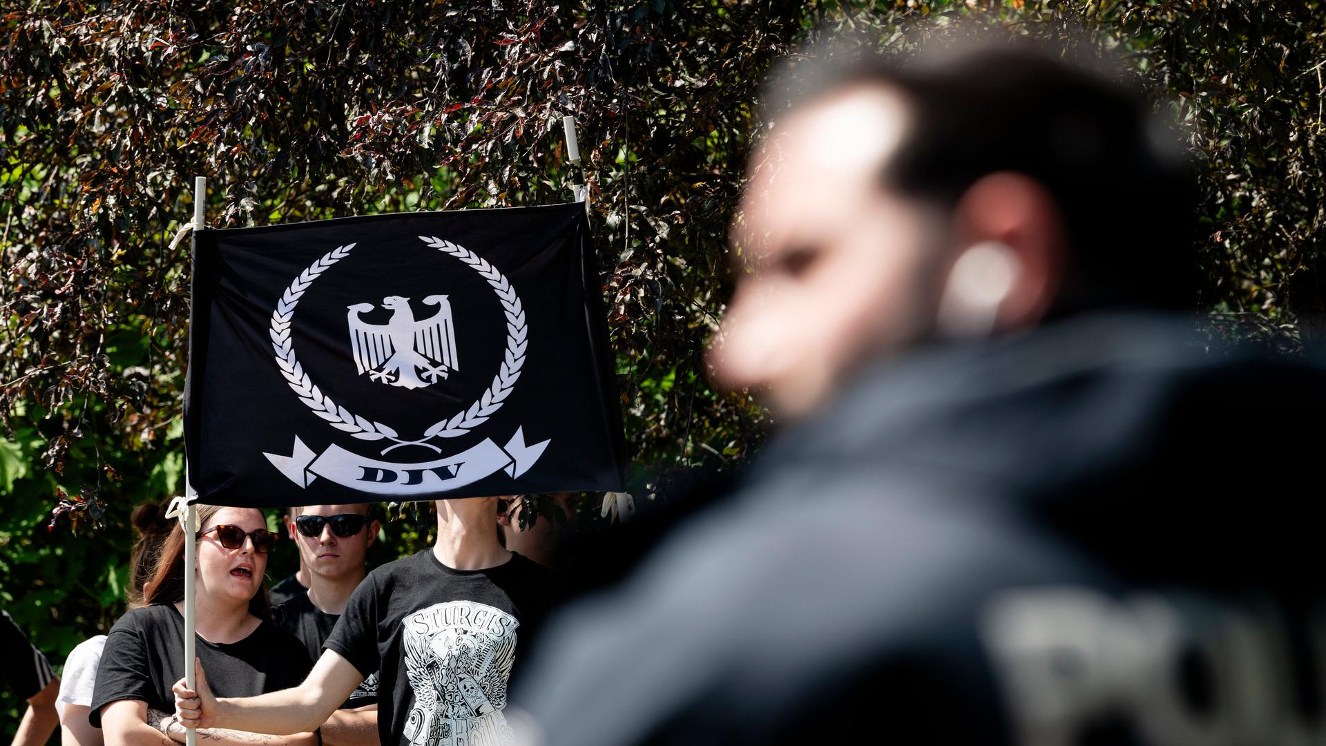 Junge Menschen tragen eine Fahne mit dem Logo der Deutschen Jugend Voran (DJV), ein weißer Adler auf schwarzem Grund, umringt von einem Eichenkranz, im Vordergrund eine verschwommene Person mit einem schwarzen Hoodie