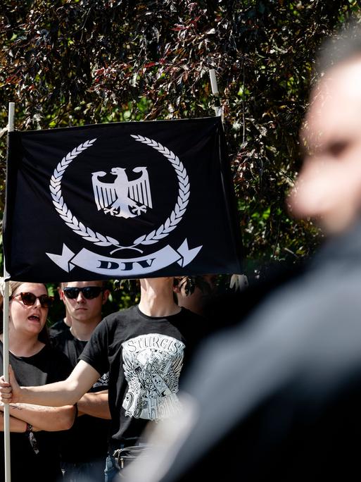 Junge Menschen tragen eine Fahne mit dem Logo der Deutschen Jugend Voran (DJV), ein weißer Adler auf schwarzem Grund, umringt von einem Eichenkranz, im Vordergrund eine verschwommene Person mit einem schwarzen Hoodie