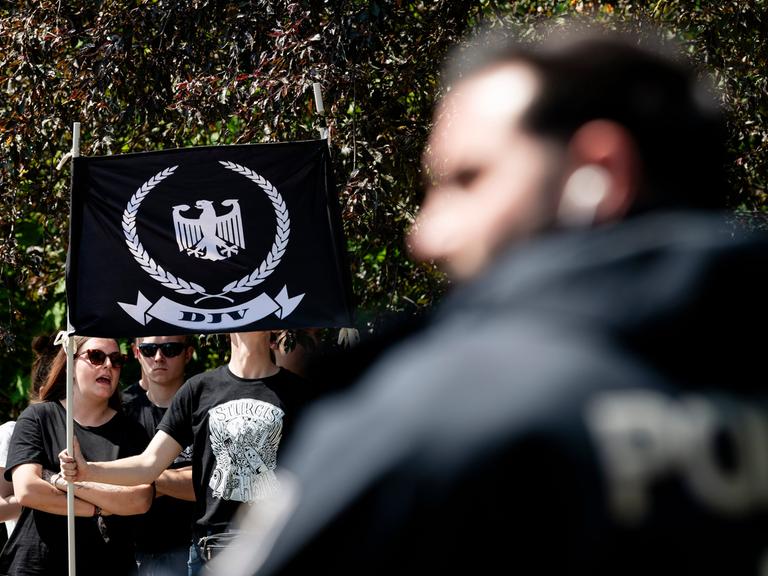 Junge Menschen tragen eine Fahne mit dem Logo der Deutschen Jugend Voran (DJV), ein weißer Adler auf schwarzem Grund, umringt von einem Eichenkranz, im Vordergrund eine verschwommene Person mit einem schwarzen Hoodie
