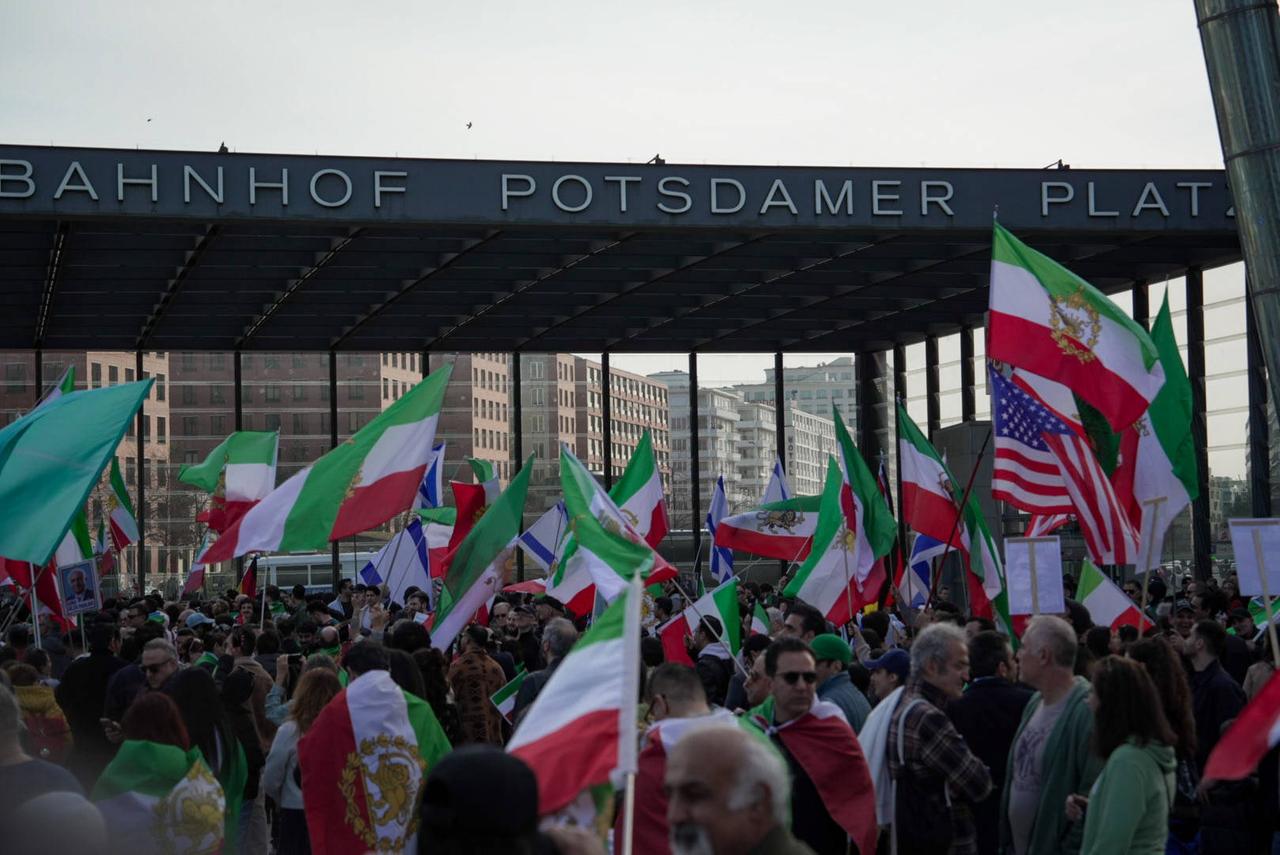 Vor einem Gebäude mit der Aufschrift "Potsdamer Platz" schwenkt eine Menschenmenge Fahnen in den Farben grün, weiß und rot.