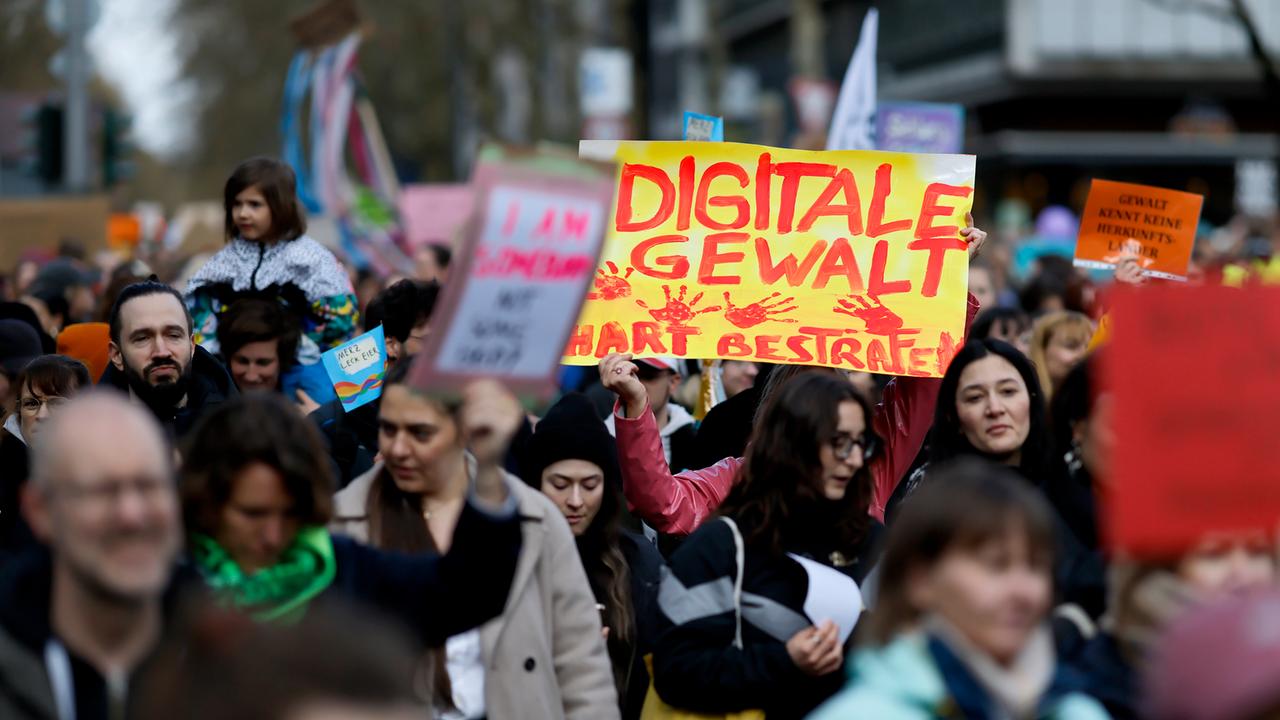 Demonstranten stehen dicht gedrängt auf der Straße und halten Schilder in die Höhe. Auf einem Schild steht "Digitale Gewalt hart bestrafen" in Rot auf Gelb.