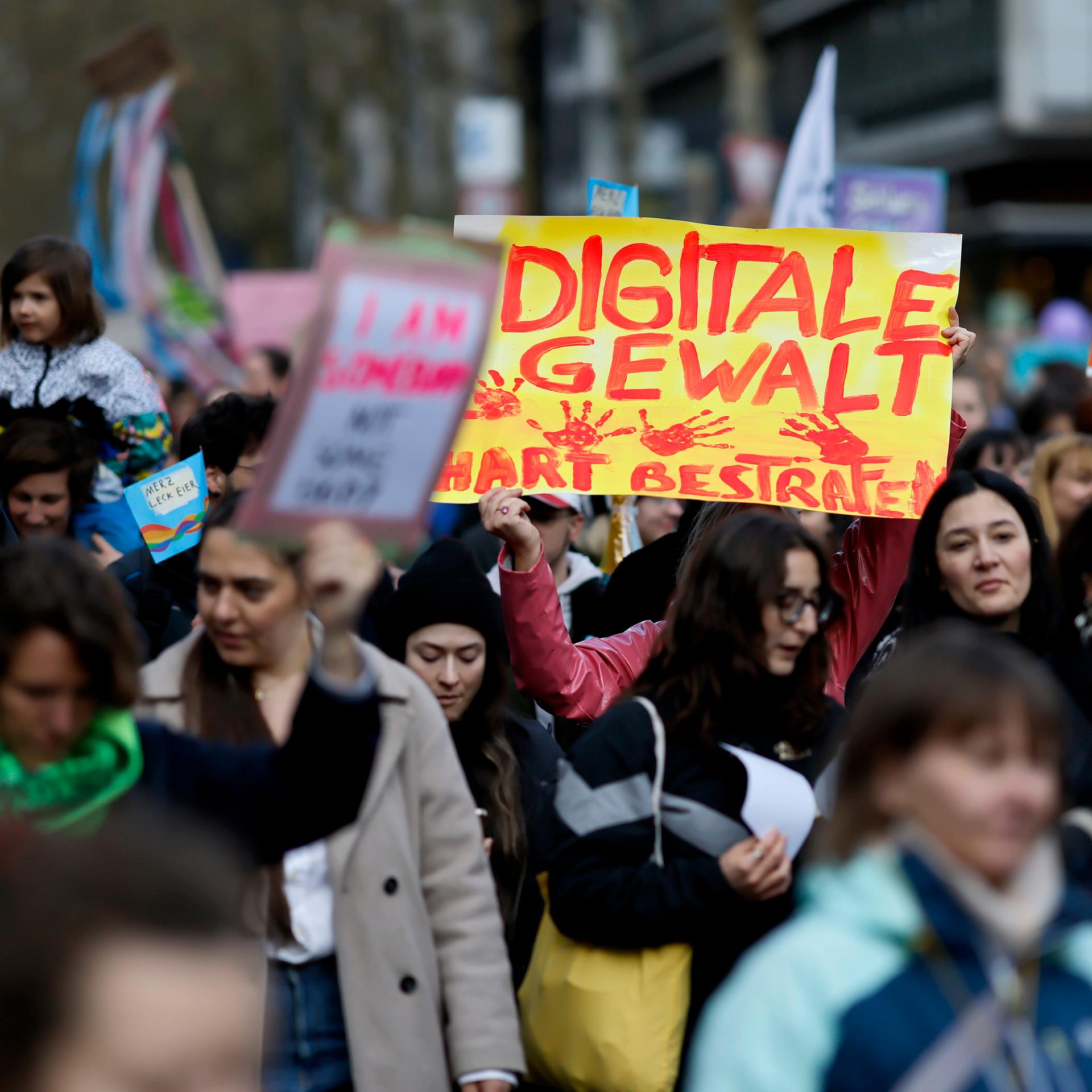 Demonstranten stehen dicht gedrängt auf der Straße und halten Schilder in die Höhe. Auf einem Schild steht "Digitale Gewalt hart bestrafen" in Rot auf Gelb.