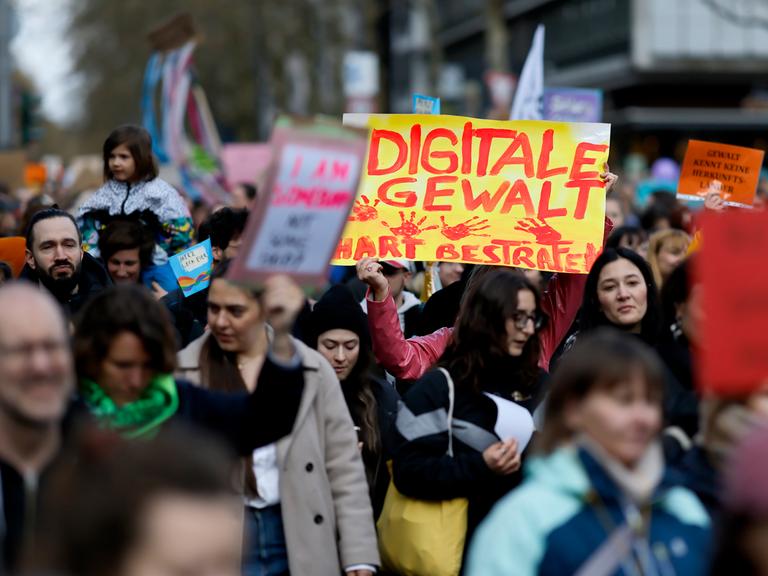 Demonstranten stehen dicht gedrängt auf der Straße und halten Schilder in die Höhe. Auf einem Schild steht "Digitale Gewalt hart bestrafen" in Rot auf Gelb.