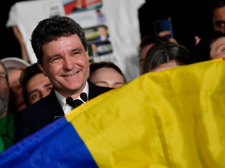 Der Kandidat Nicusor Dan spricht zu Journalisten nach dem Ende der Abstimmung. Vor ihm ist eine rumänische Flagge, hinter ihm stehen seine Anhänger. Er lächelt. Der Kandidat Nicusor Dan spricht zu Journalisten nach dem Ende der Abstimmung. Vor ihm ist eine rumänische Flagge, hinter ihm stehen seine Anhänger. Er lächelt.