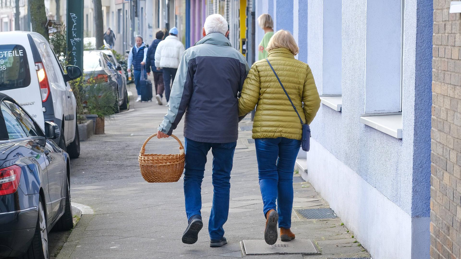 Ein älteres Paar läuft mit einem Einkaufskorb einen Bürgersteig entlang. Man sieht sie von hinten.