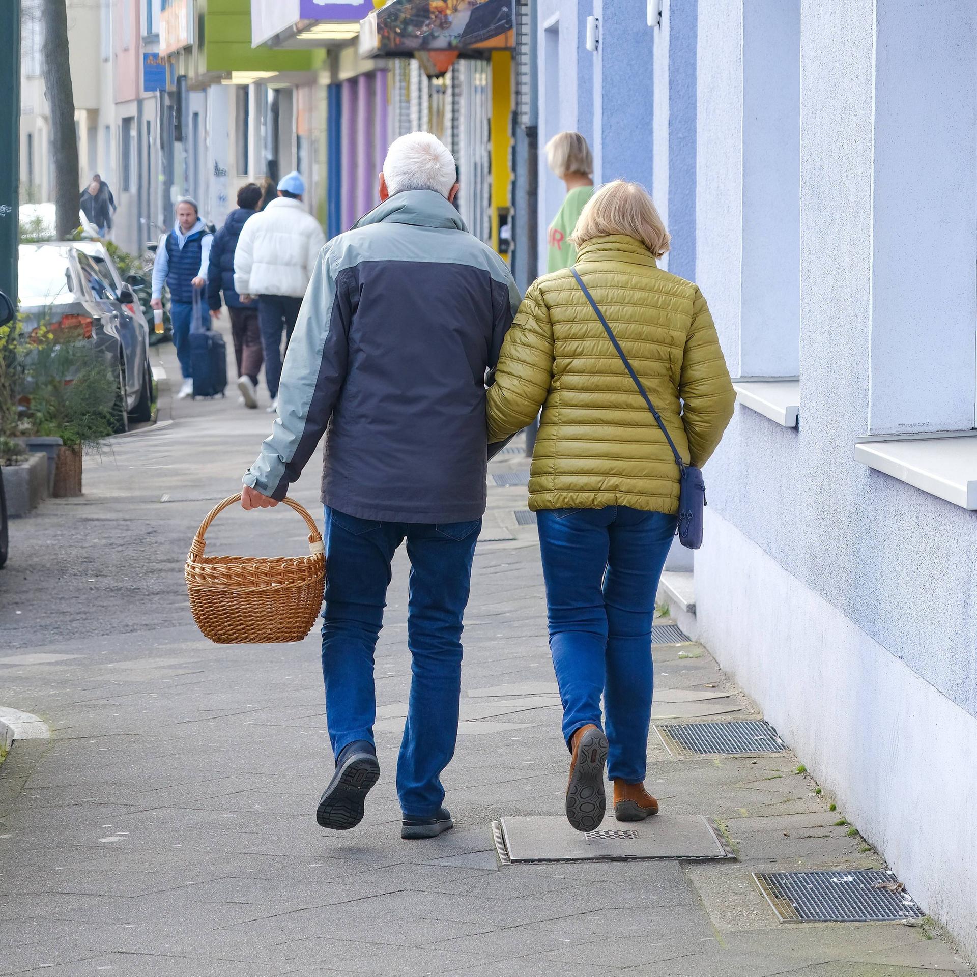 Ein älteres Paar läuft mit einem Einkaufskorb eine Straße entlang.