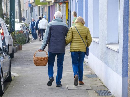Ein älteres Paar läuft mit einem Einkaufskorb eine Straße entlang.