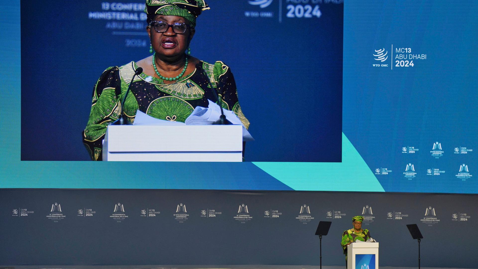Die Generaldirektorin der Welthandelsorganisation, Ngozi Okonjo-Iweala, spricht beim 13. Ministertreffen der Welthandelsorganisation. Man sieht sie einmal klein an einem Rednerpult und einmal im Hintergrund groß auf einer Leinwand. Die Generaldirektorin der Welthandelsorganisation, Ngozi Okonjo-Iweala, spricht beim 13. Ministertreffen der Welthandelsorganisation. Man sieht sie einmal klein an einem Rednerpult und einmal im Hintergrund groß auf einer Leinwand.