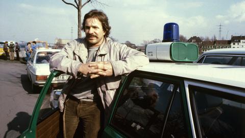 Götz George zu Beginn der Dreharbeiten zu dem WDR-Tatort "Duisburg-Ruhrort" am in Duisburg. Er trägt Parka und Schnauzer und steht lässig an ein Polizeiauto gelehnt, im Hintergrund ist ein weiteres Auto zu sehen. 