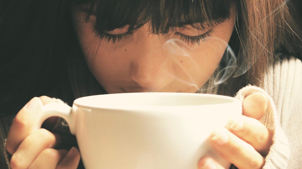 Eine junge Frau hält mit beiden Händen eine weiße Tasse in den Händen, aus der Dampf aufsteigt.