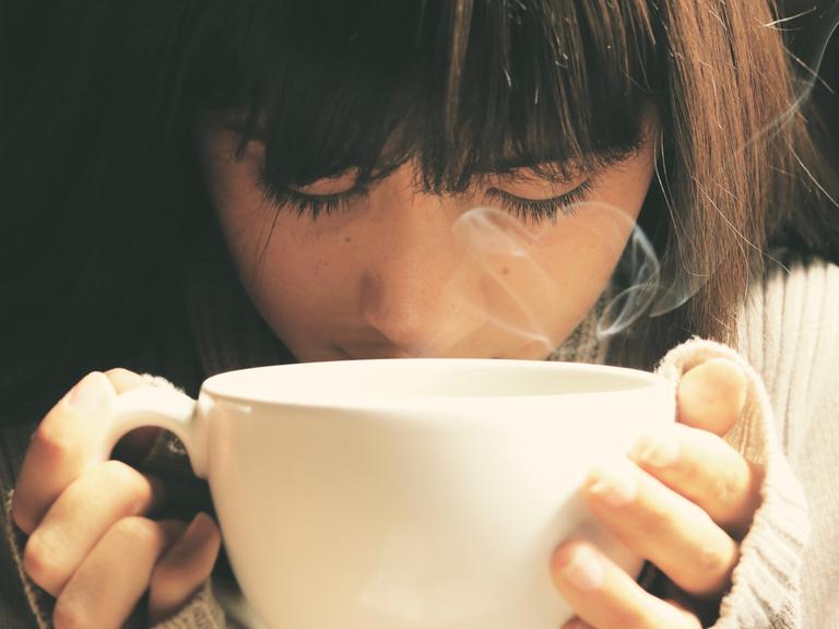 Eine junge Frau hält mit beiden Händen eine Tasse Tee in den Händen, aus der zufällig Dampf in Herzform aufsteigt.