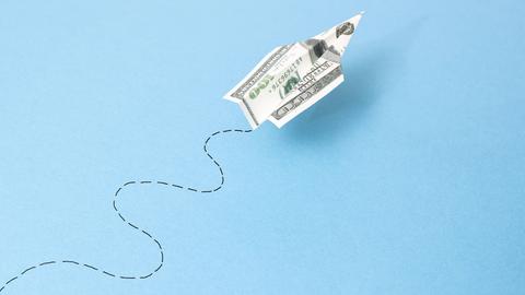 Illustration: Ein Papierflugzeug aus einem 100 Dollar-Geldschein fliegt auf blauem Hintergrund eine Zickzacklinie.