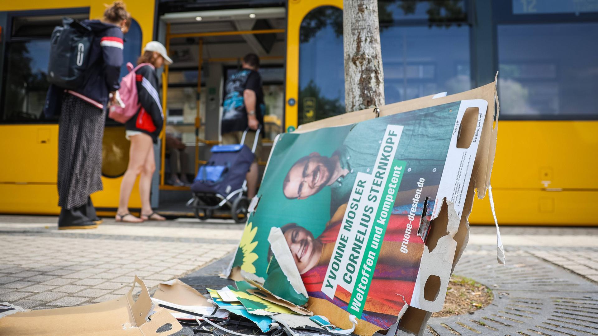 Zerfetzte und heruntergerissene Wahlplakate der Grünen liegen auf dem Boden. im Hintergrund steigen Menschen in eine Straßenbahn ein.