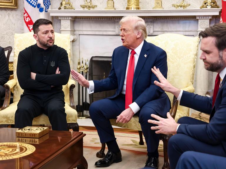 Eine Pressekonferenz im Weißen Haus in Washington. Links auf einem Sessel sitzt der ukrainische Präsident Wolodymyr Selenskyj mit verschränkten Armen, rechts von ihm gestikulieren Präsident Donald Trump und Vize-Präsident J. D. Vance mit erhobenen Händen.