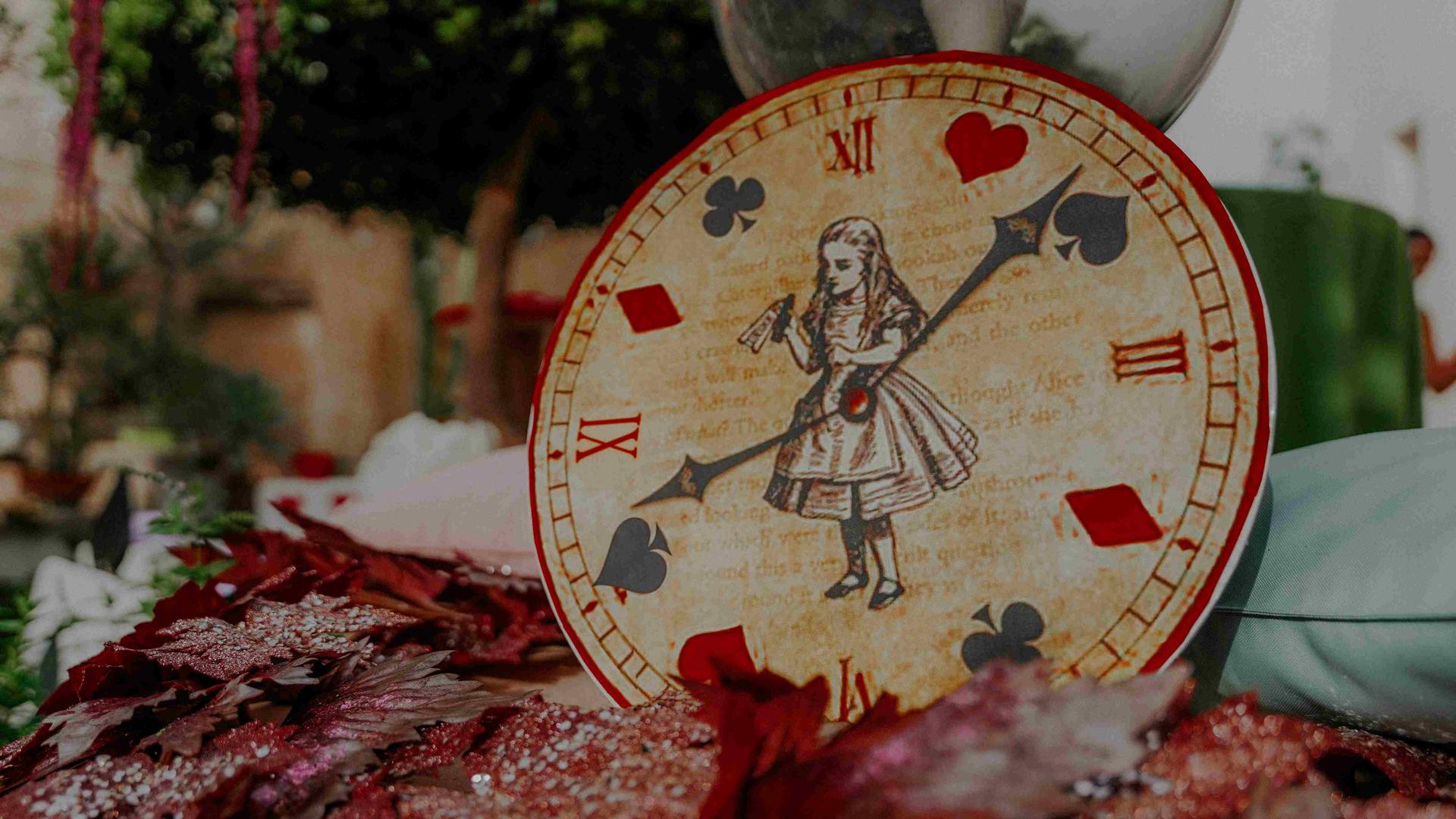 Eine Uhr, auf deren Ziffernblatt Alice im Wunderland zu sehen ist, und die Zeiger zeigen auf Pik, Herz, Karo und Kreuz.