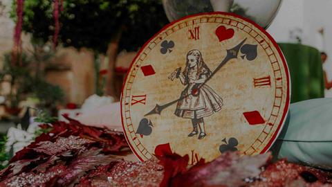 Eine Uhr, auf deren Ziffernblatt Alice im Wunderland zu sehen ist, und die Zeiger zeigen auf Pik, Herz, Karo und Kreuz.