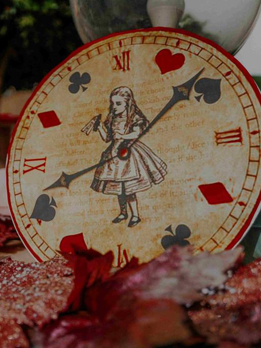 Eine Uhr, auf deren Ziffernblatt Alice im Wunderland zu sehen ist, und die Zeiger zeigen auf Pik, Herz, Karo und Kreuz.