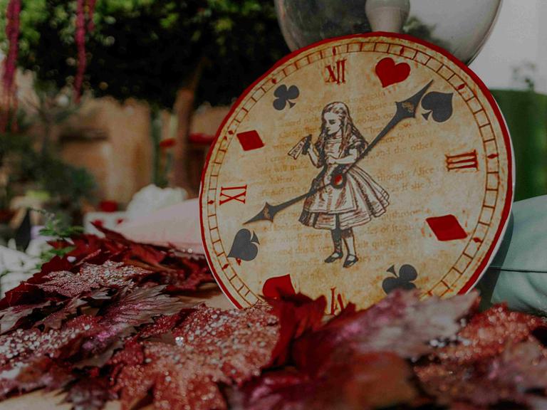 Eine Uhr, auf deren Ziffernblatt Alice im Wunderland zu sehen ist, und die Zeiger zeigen auf Pik, Herz, Karo und Kreuz.